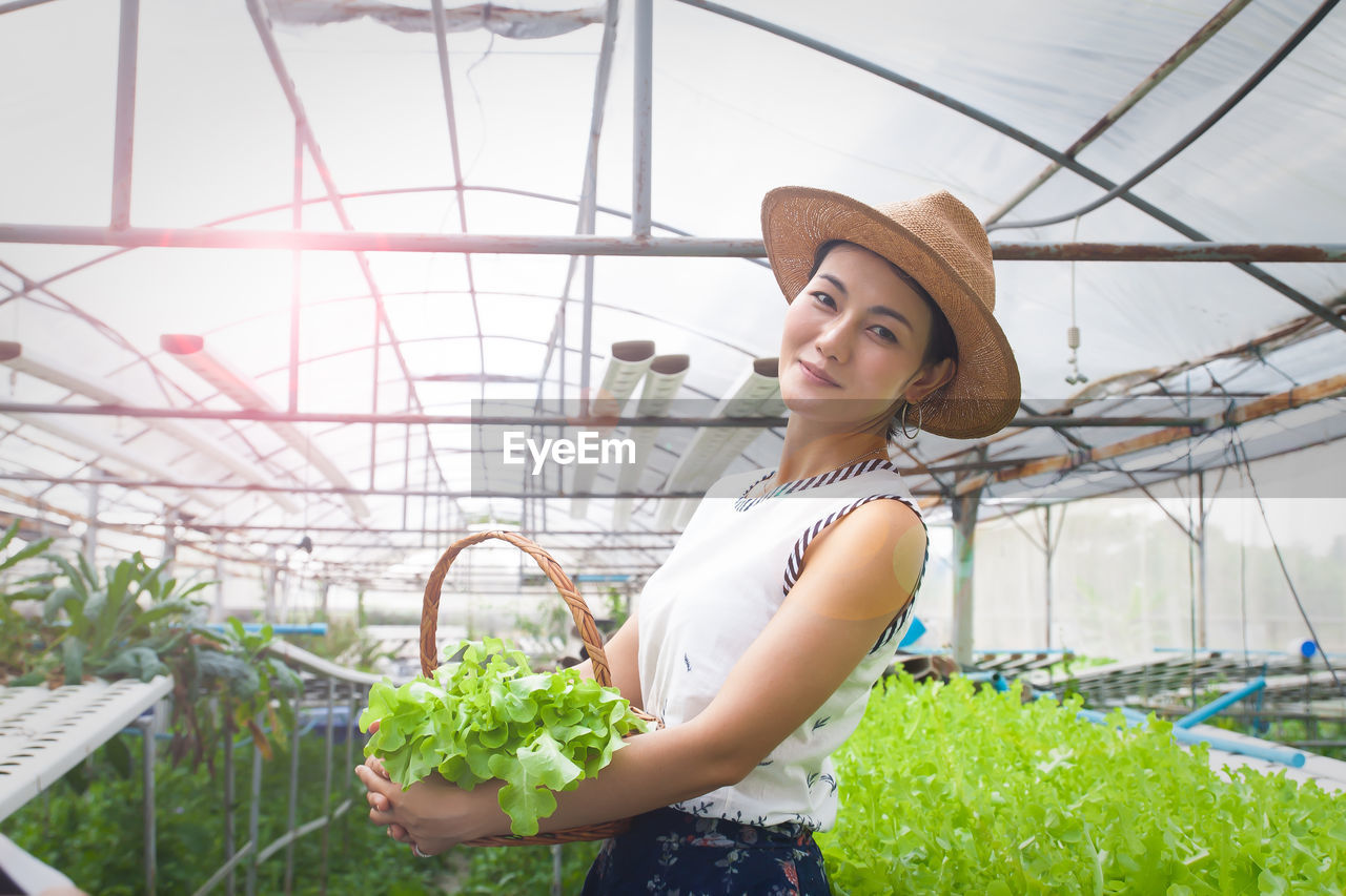 Portrait of woman holding vegetables while standing in greenhouse