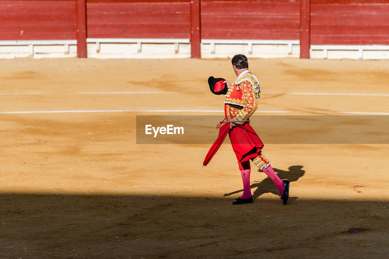 Back view of unrecognizable bullfighter in fancy costume taking off hat after corrida performance while standing on sandy arena