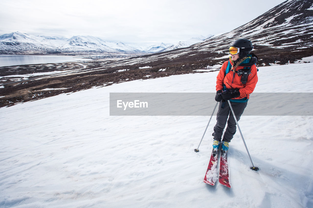 A woman standing with skis on snow with mountains and water behind