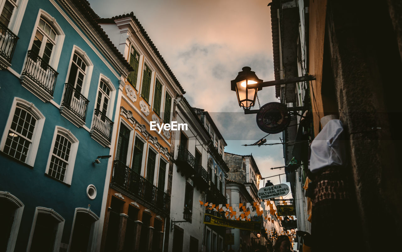 LOW ANGLE VIEW OF ILLUMINATED STREET AMIDST BUILDINGS AGAINST SKY