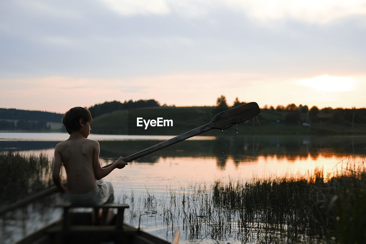 MAN SITTING ON LAKE AGAINST SKY AT SUNSET