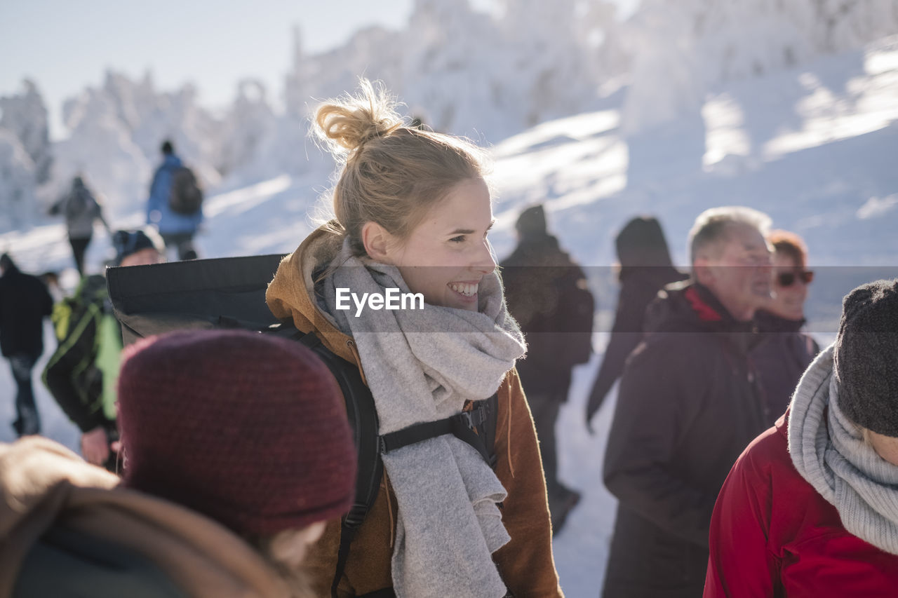 Smiling young woman wearing warm clothing while looking away by people in winter