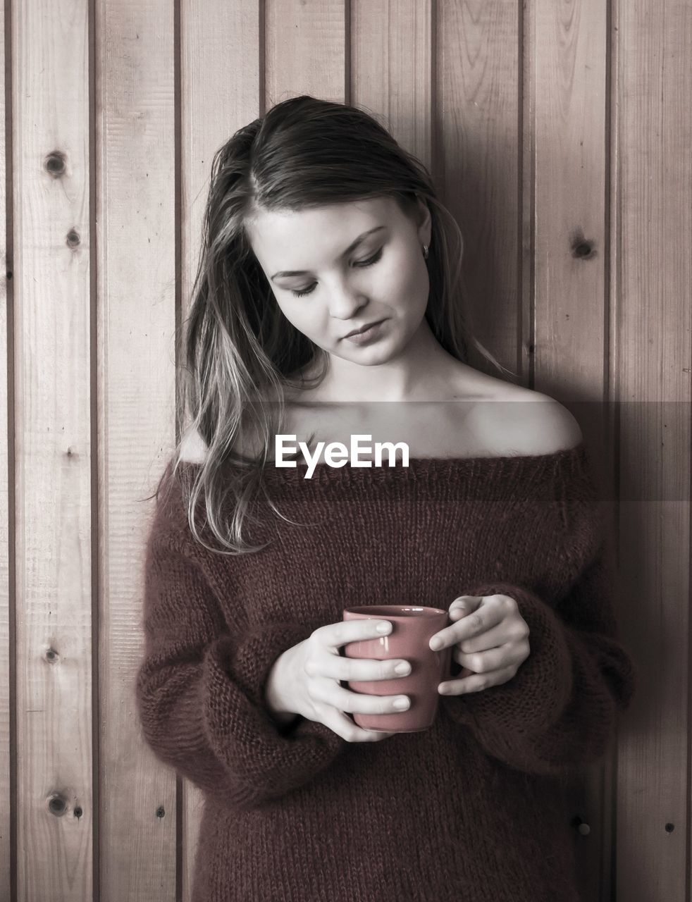 Thoughtful young woman holding coffee cup against wooden wall