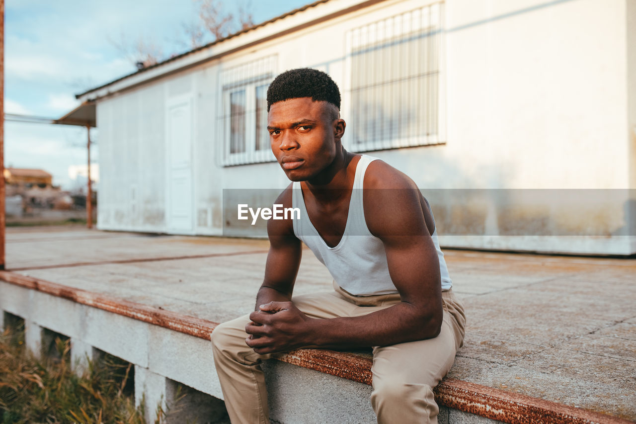 Young masculine african american male in undershirt with clasped hands looking at camera against house