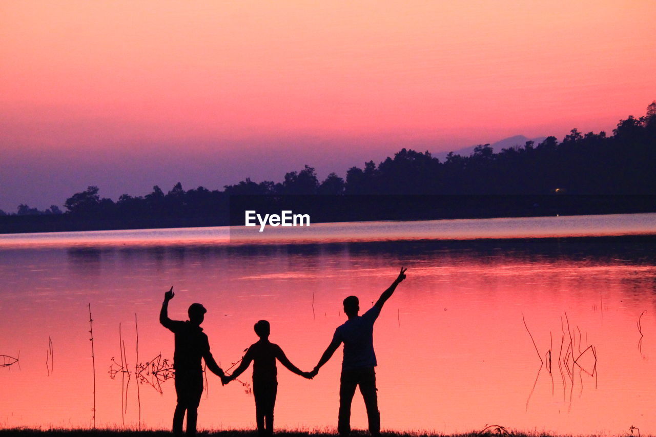 Rear view of silhouette friends standing by river during sunset