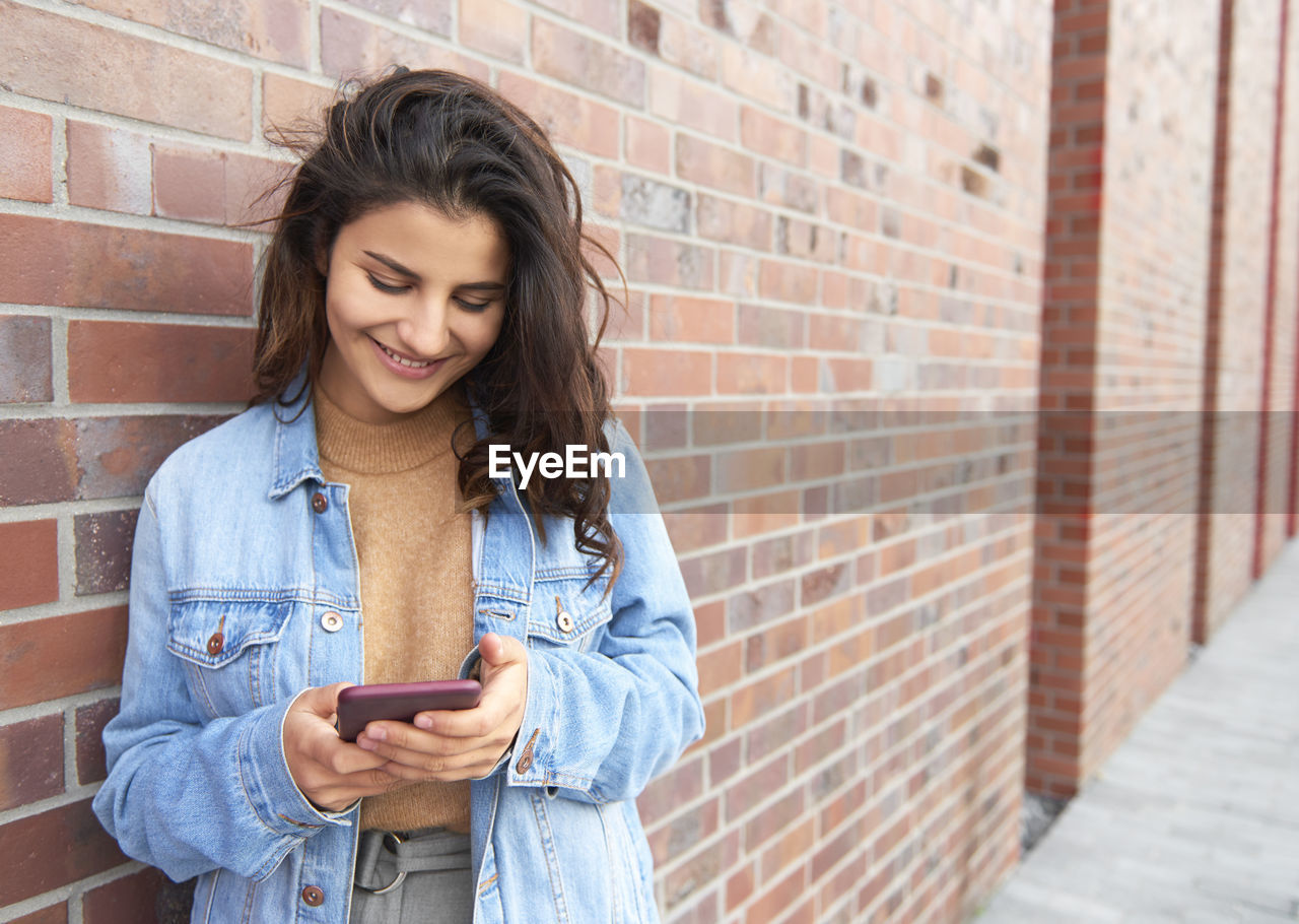 Smiling young woman using mobile phone against wall