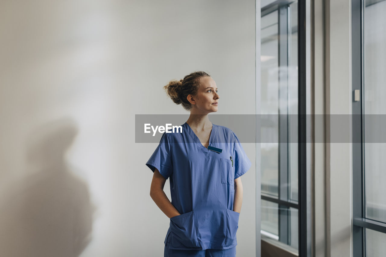 Thoughtful young nurse standing with hands in pockets against wall at hospital