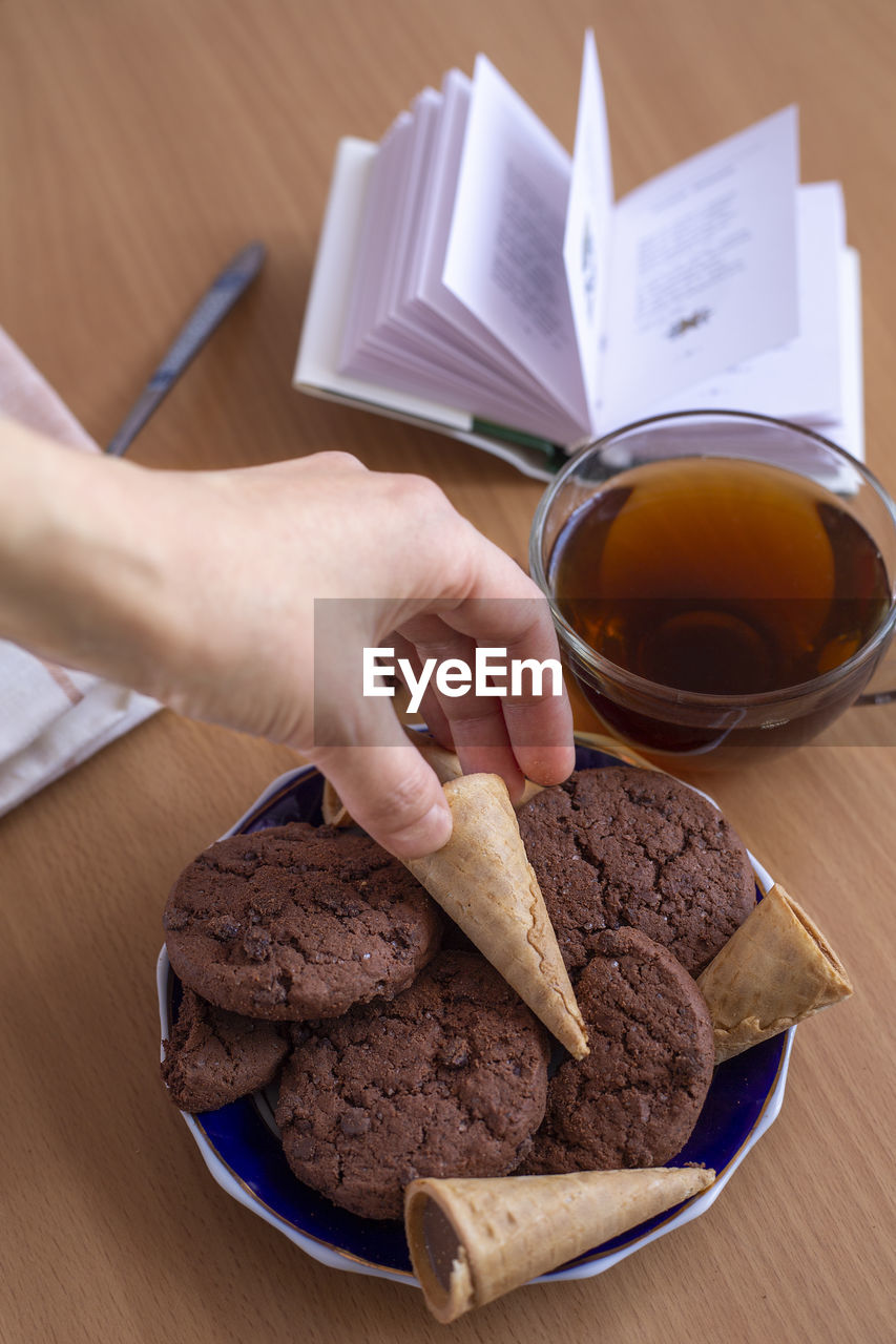 A woman's hand takes a waffle tube from a saucer with sweets, next to a cup of tea and an open book.