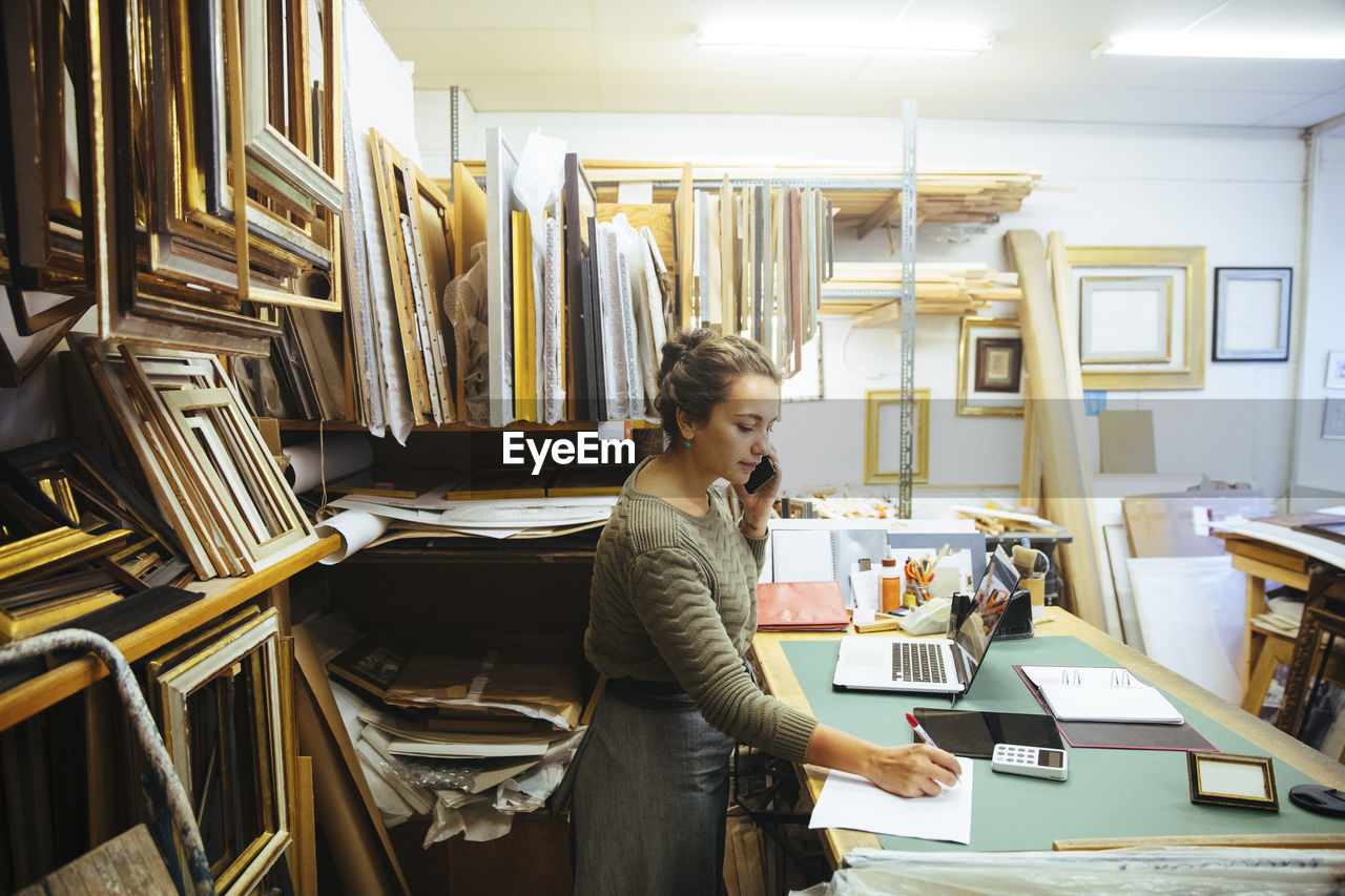 Confident young craftswoman talking on smart phone while writing in paper at workbench in framing store