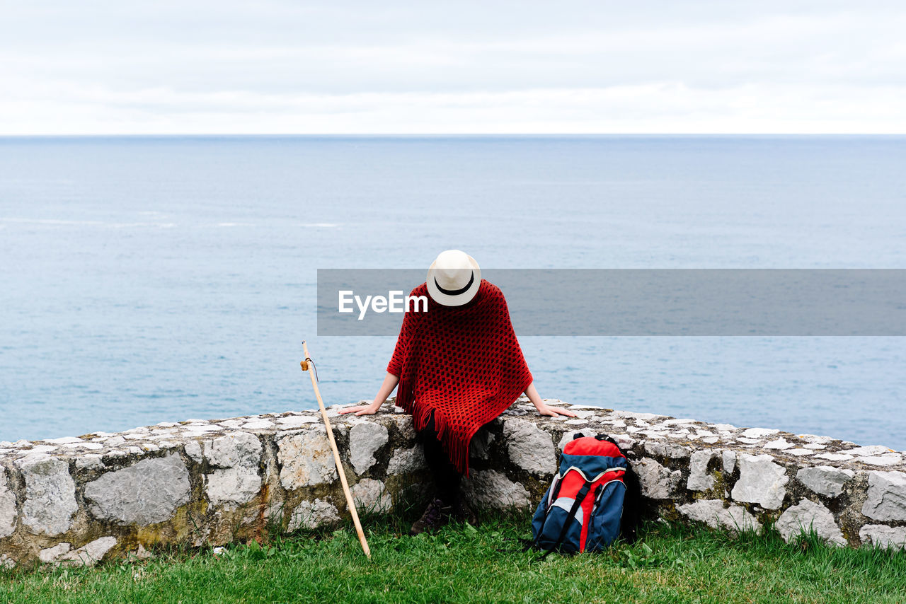 Happy young female backpacker in stylish clothes sitting on stone border against sea bay and looking down while resting during camino de santiago route in spain