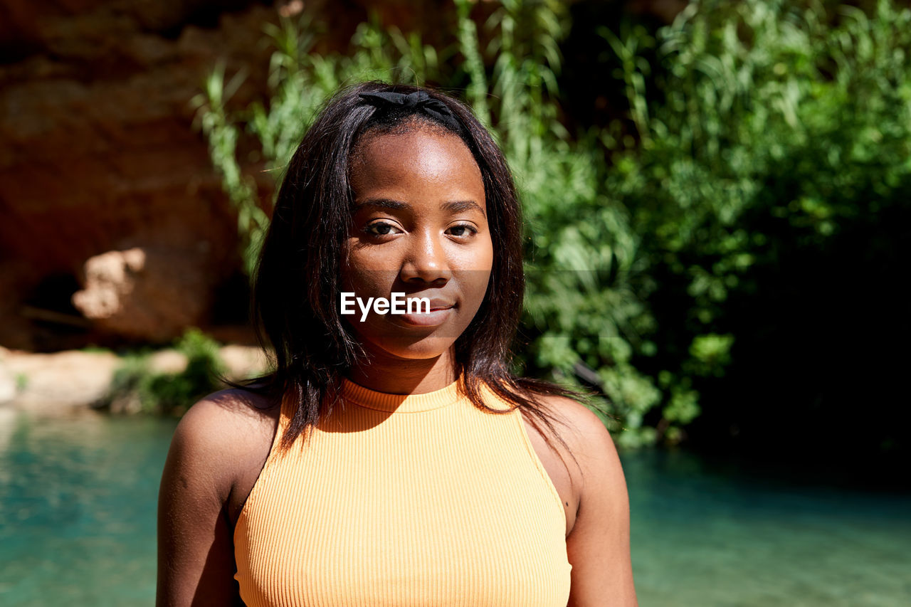 African american female traveler in casual clothes looking at camera while standing on shore of clean lake against cliff with waterfall on summer weekend day