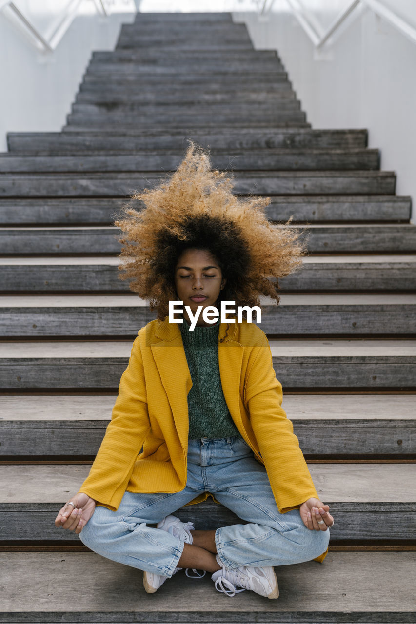 Afro woman with tousled hair meditating while sitting on staircase