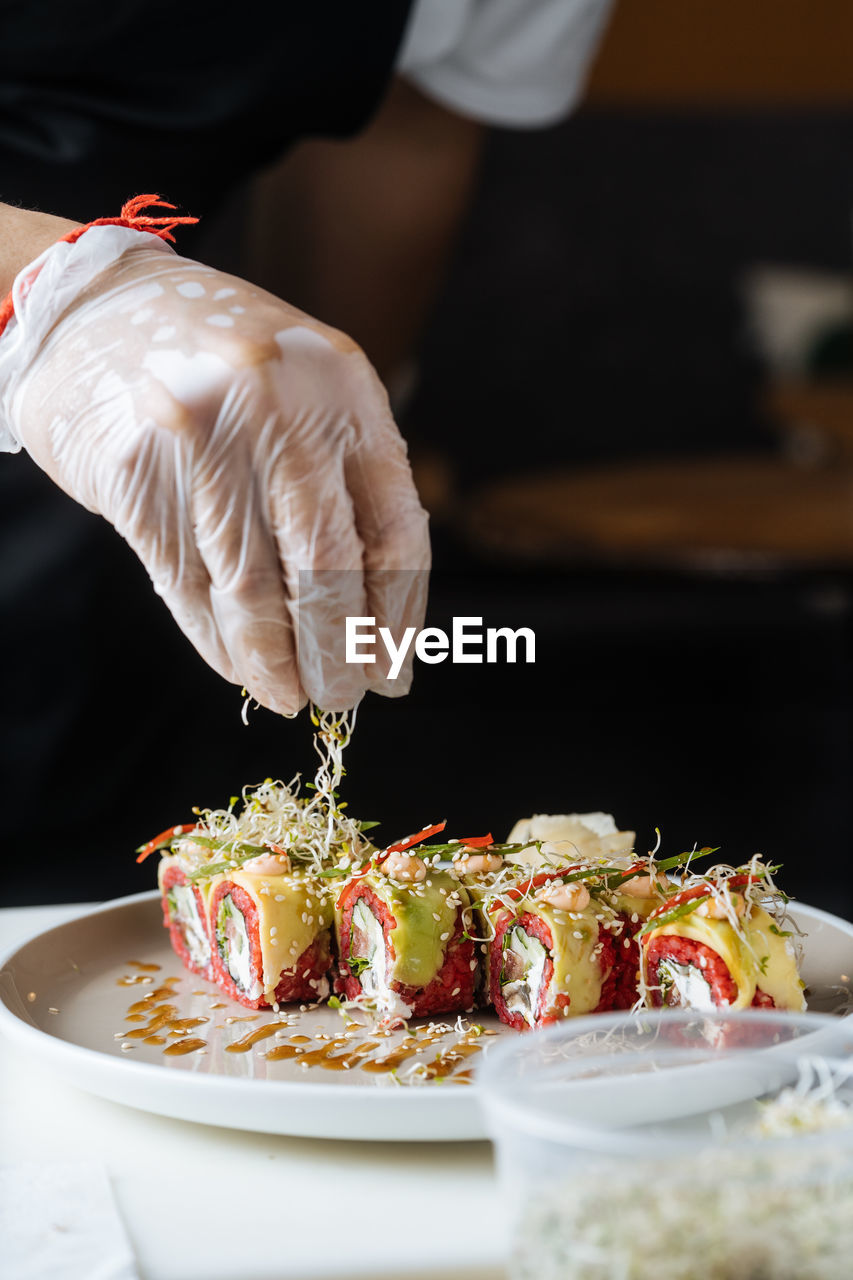 Crop cook in disposable gloves decorating fresh tasty sushi with greens on white plate on table