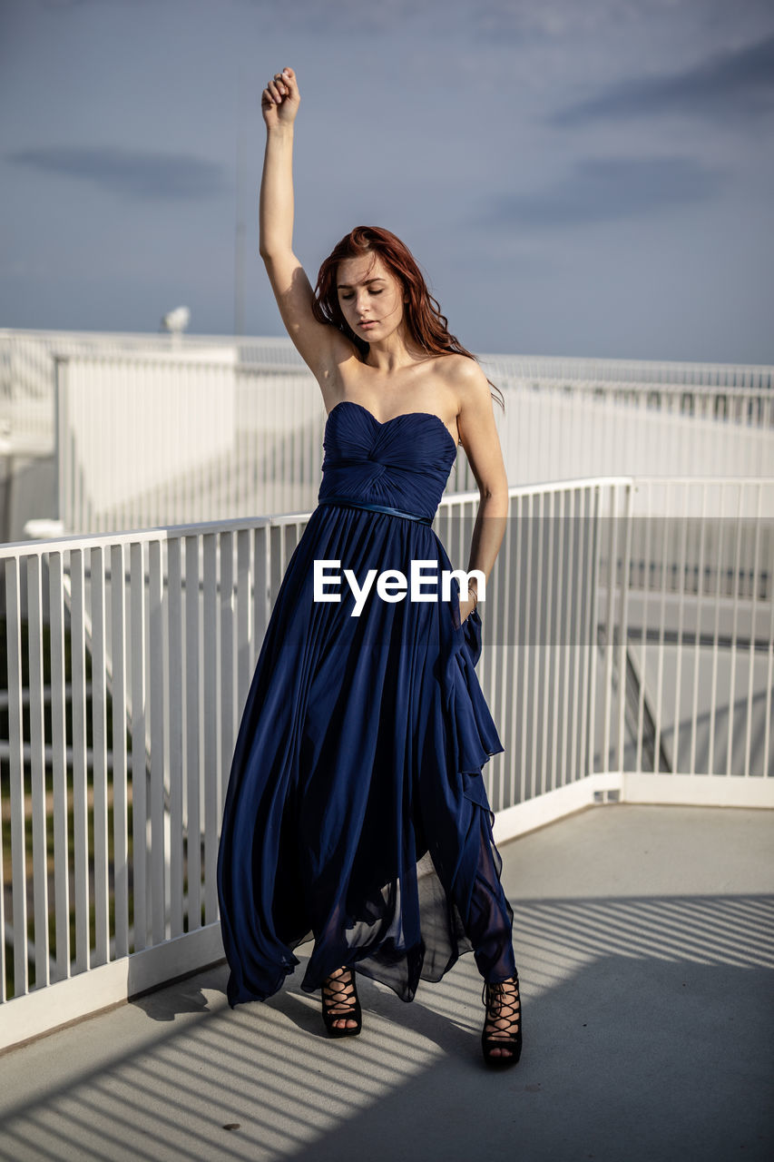 Woman in dress with hand raised standing in balcony against sky