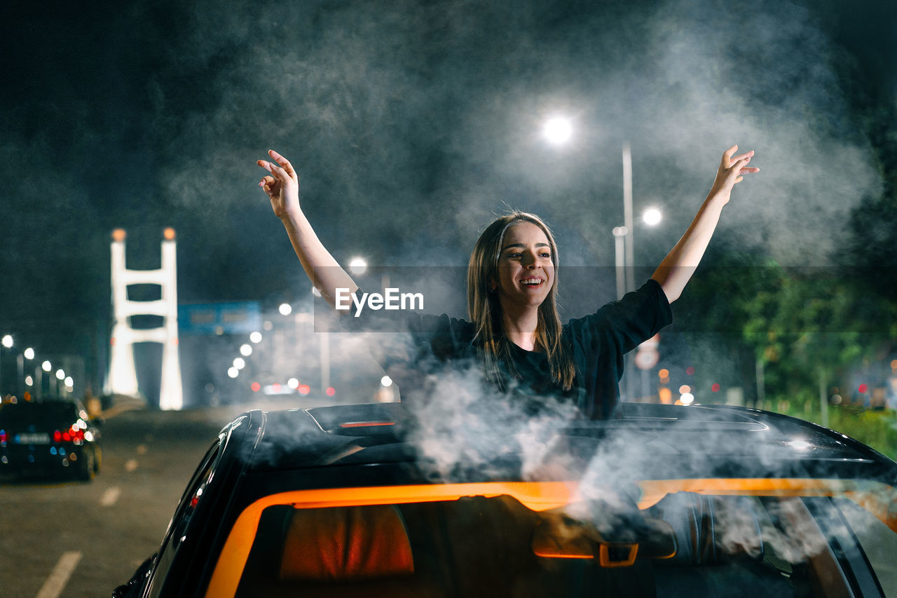 Happy young woman with arms raised through sun roof of a car in the city at night