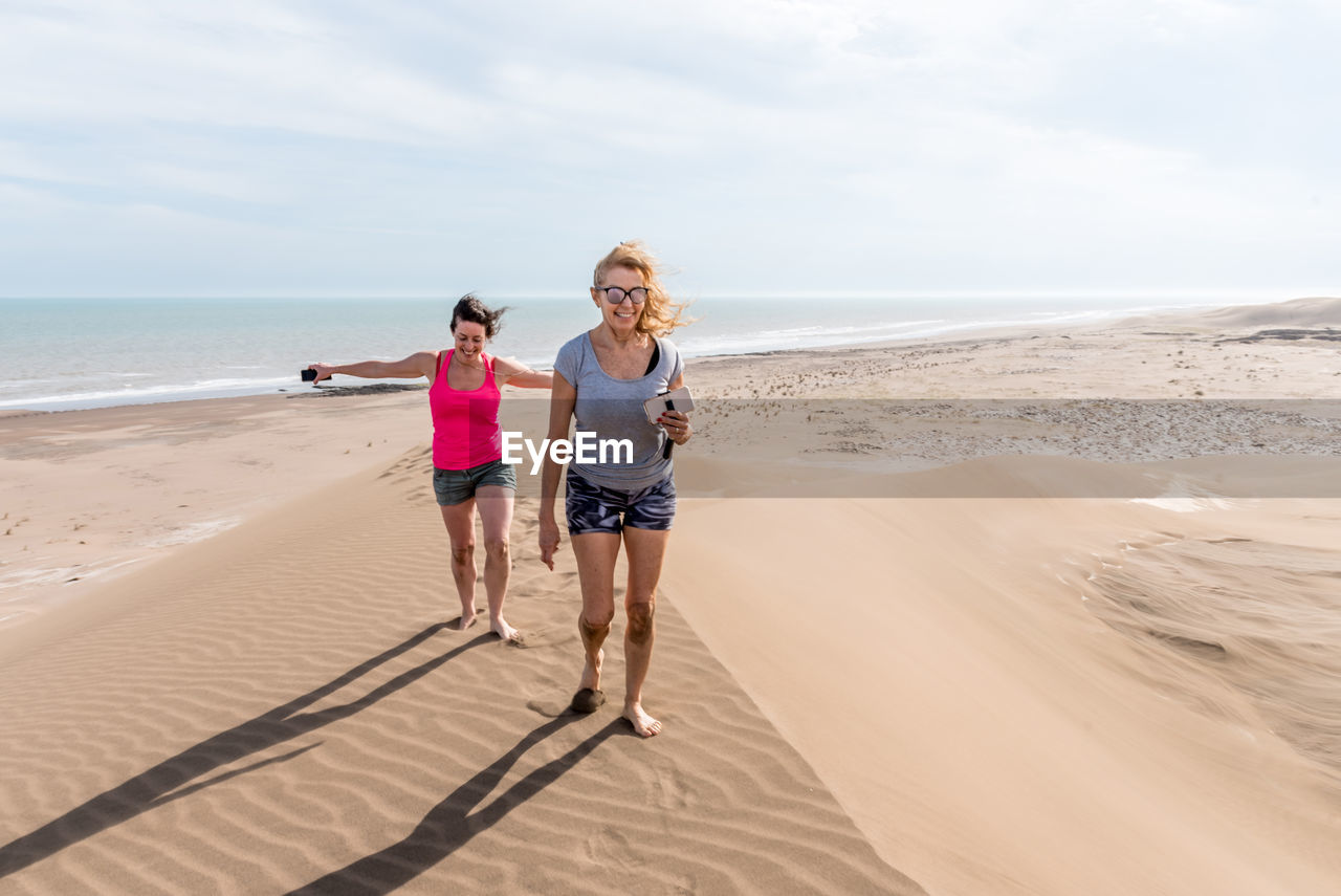Front view of two women walking on top of a large dune, one of them with arms open to the wind