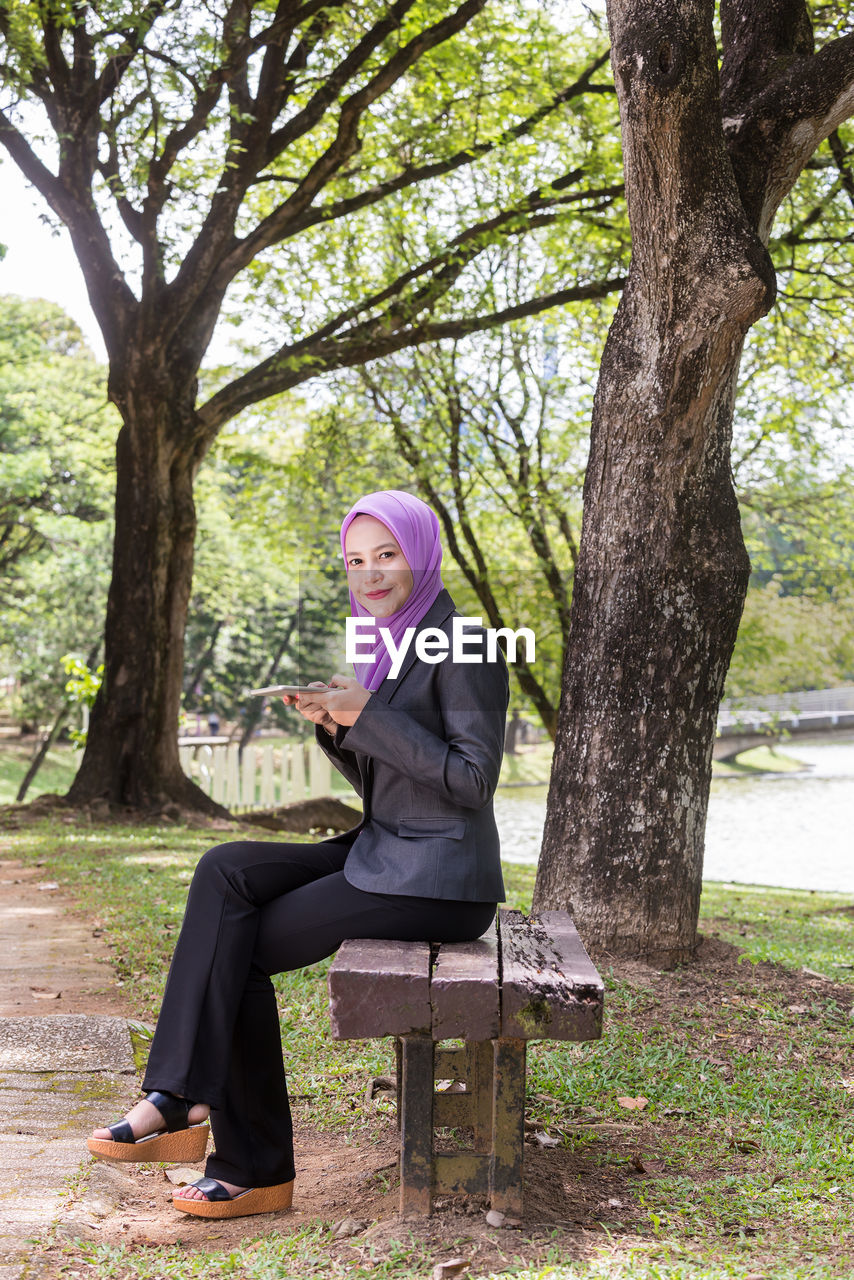 Portrait of young woman wearing hijab using mobile phone while sitting in park