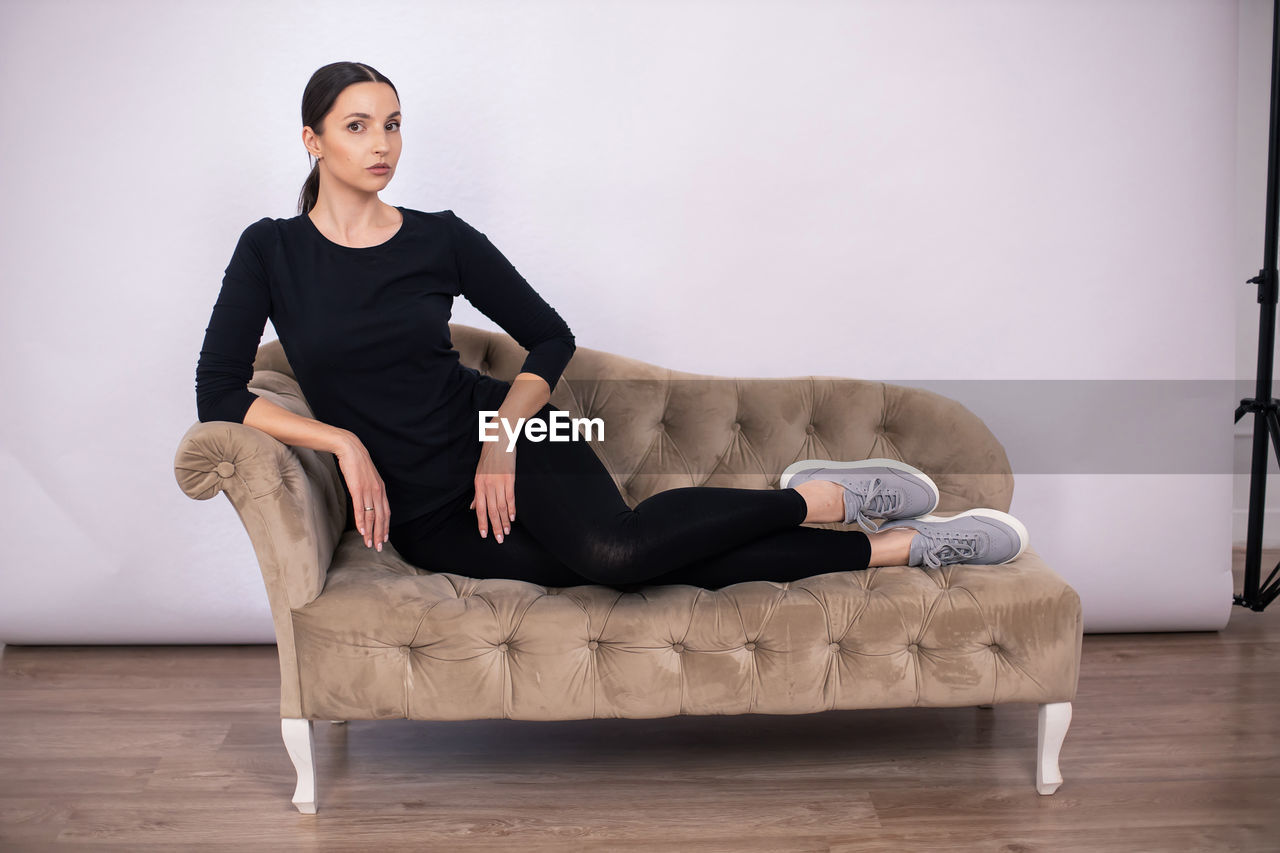 Full length woman poses on a sofa in black tight fitting clothes and sneakers in a studio on a white