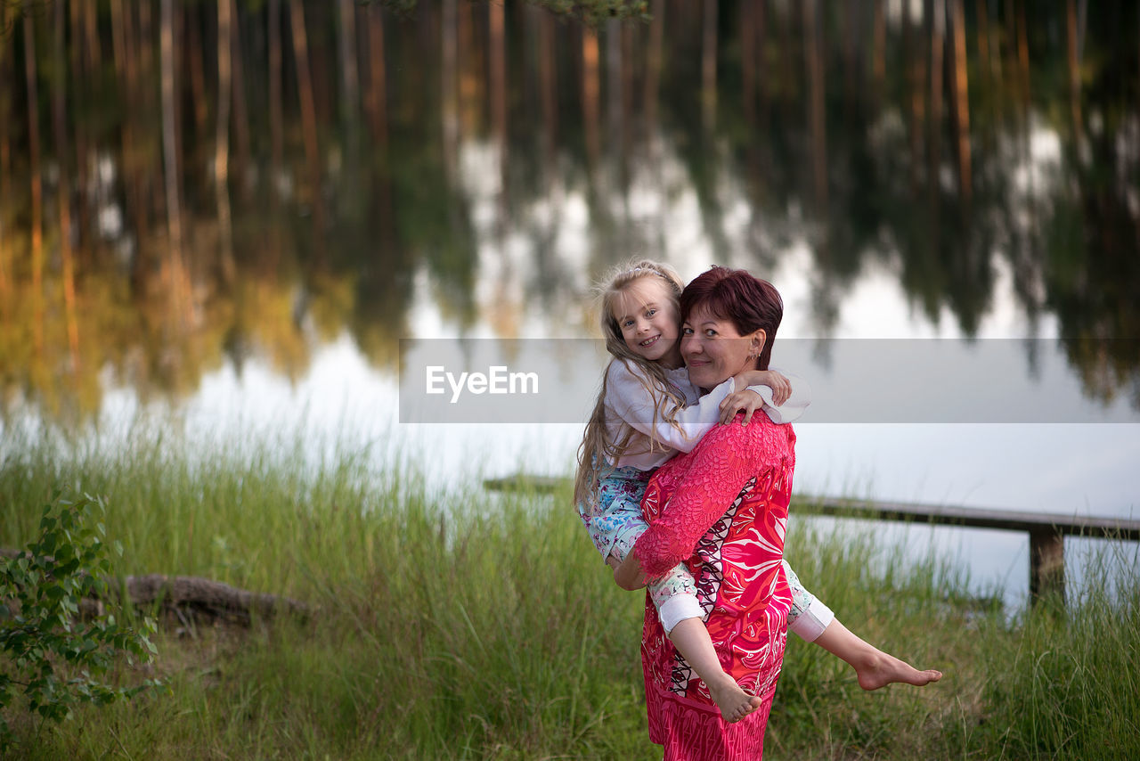 Portrait of woman carrying daughter standing on field against lake