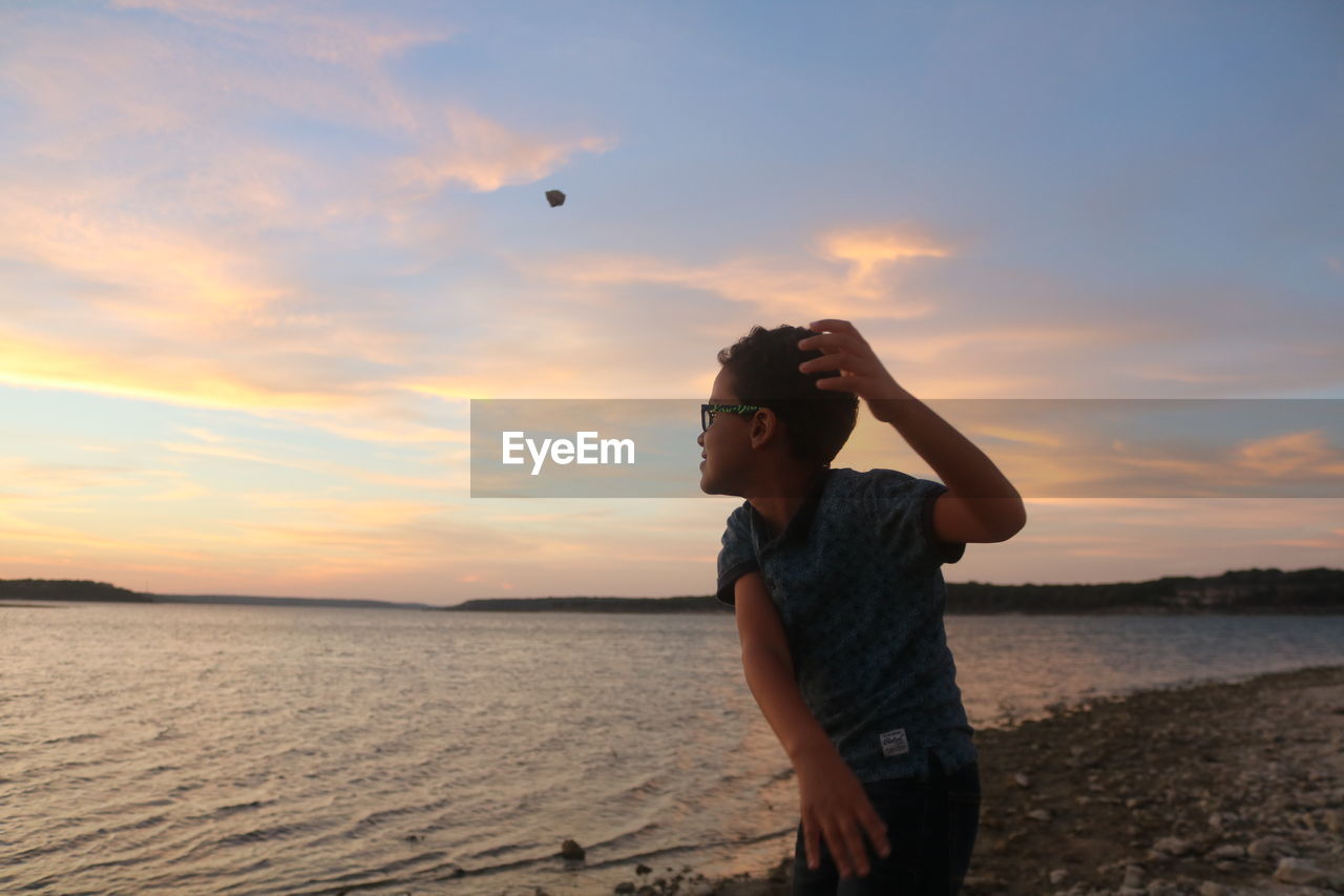 Boy throwing rock in lake against sky during sunset