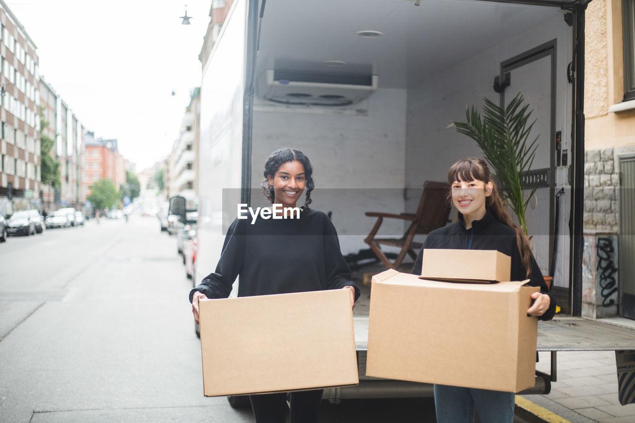 Portrait of smiling female movers unloading boxes from truck on street in city