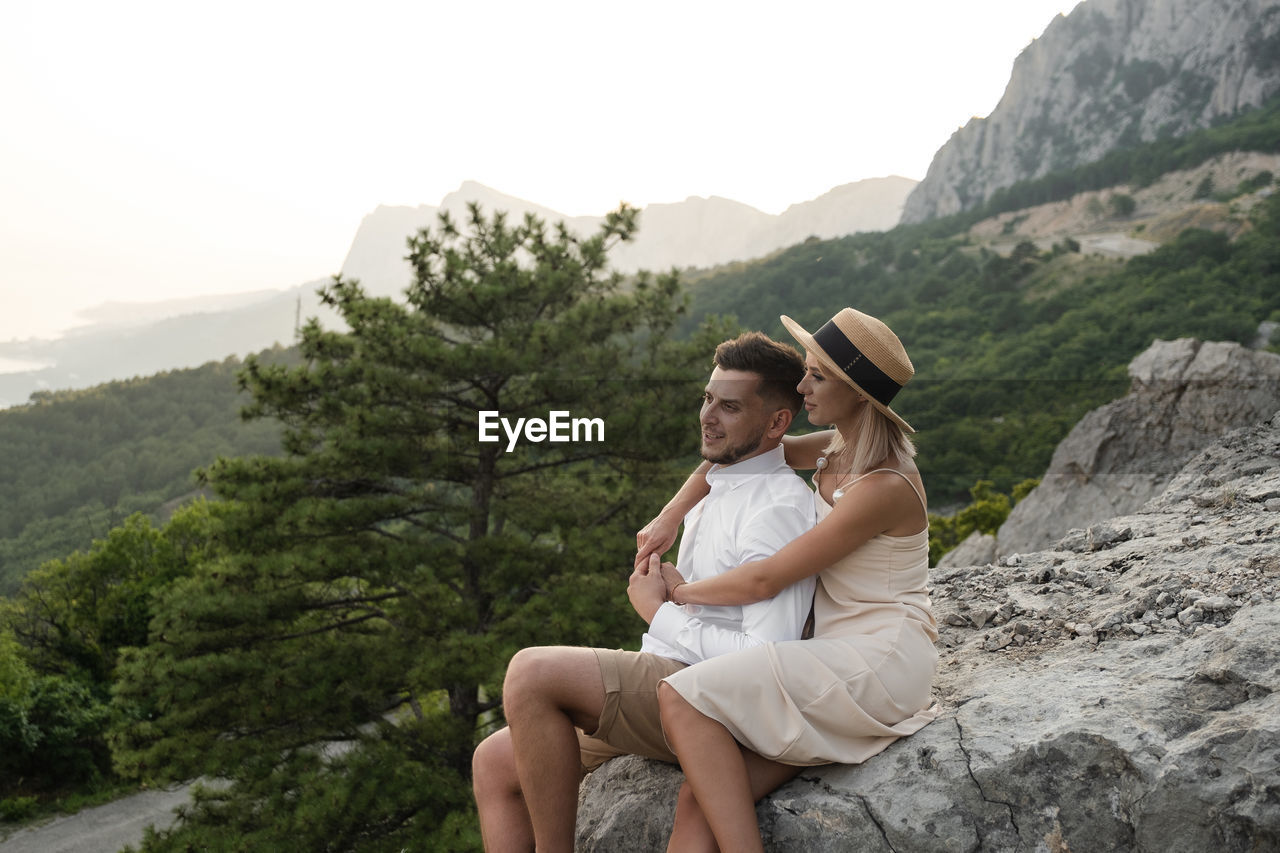 PEOPLE SITTING ON ROCK AGAINST MOUNTAIN