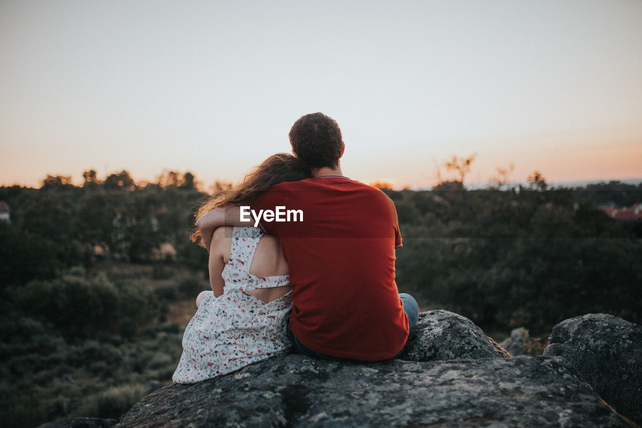 Rear view of couple sitting on rock against clear sky during sunset