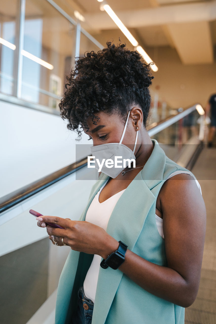 Side view of positive young african american female in casual outfit and protective mask using mobile phone while standing inside public building with escalator