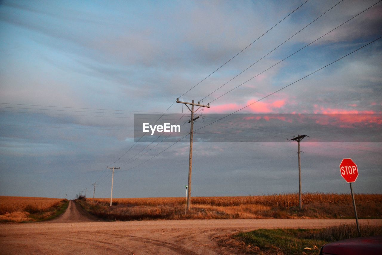 Telephone poles on landscape against cloudy sky during sunset