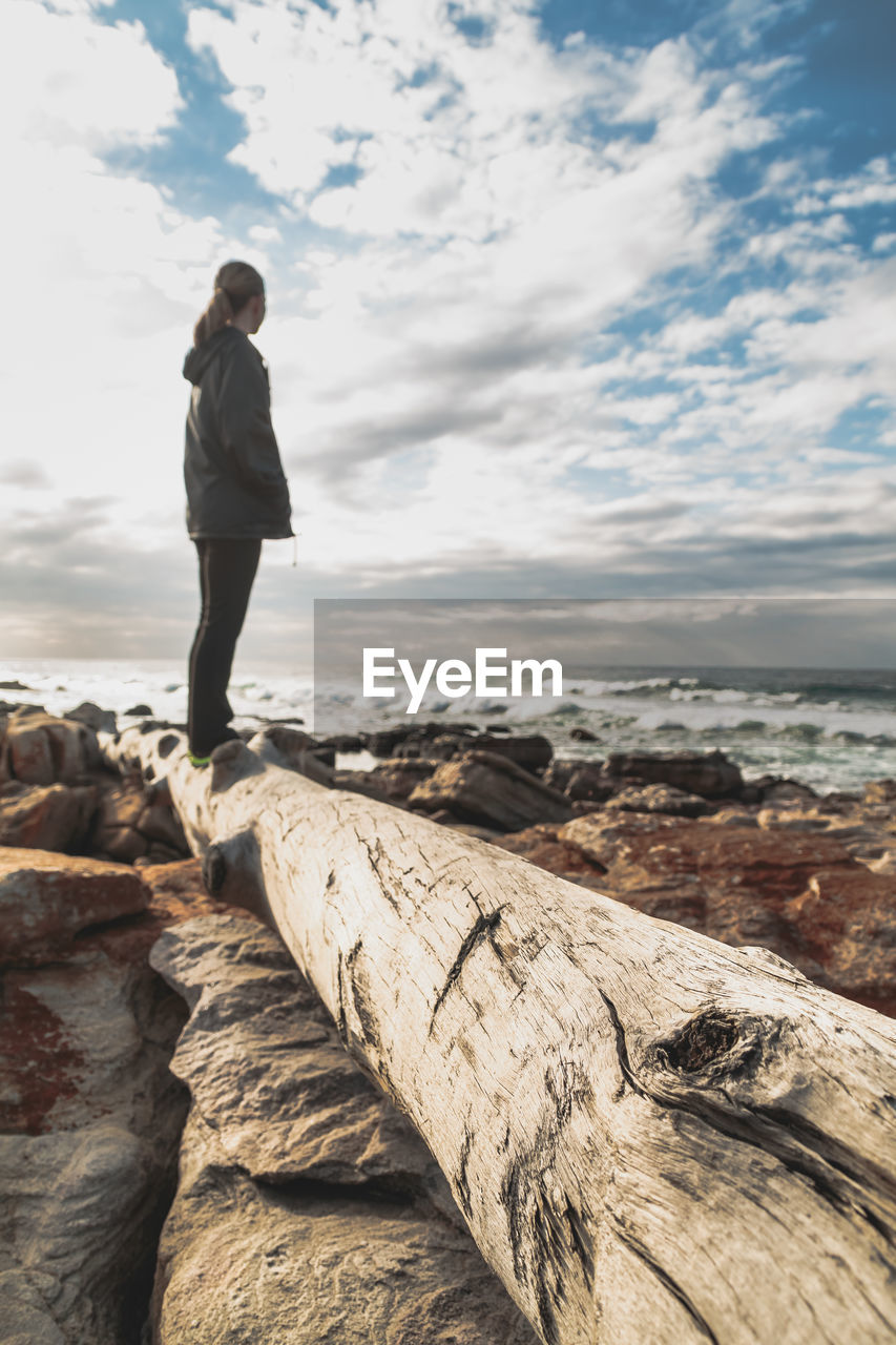 Side view of woman standing on log by sea against cloudy sky