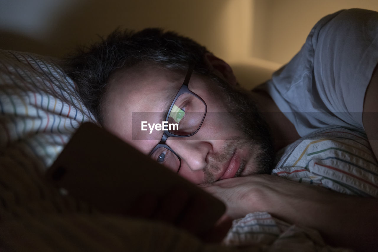 Sleepy male with glasses surfing in web before sleeping. social media addict, sleeplessness concept.