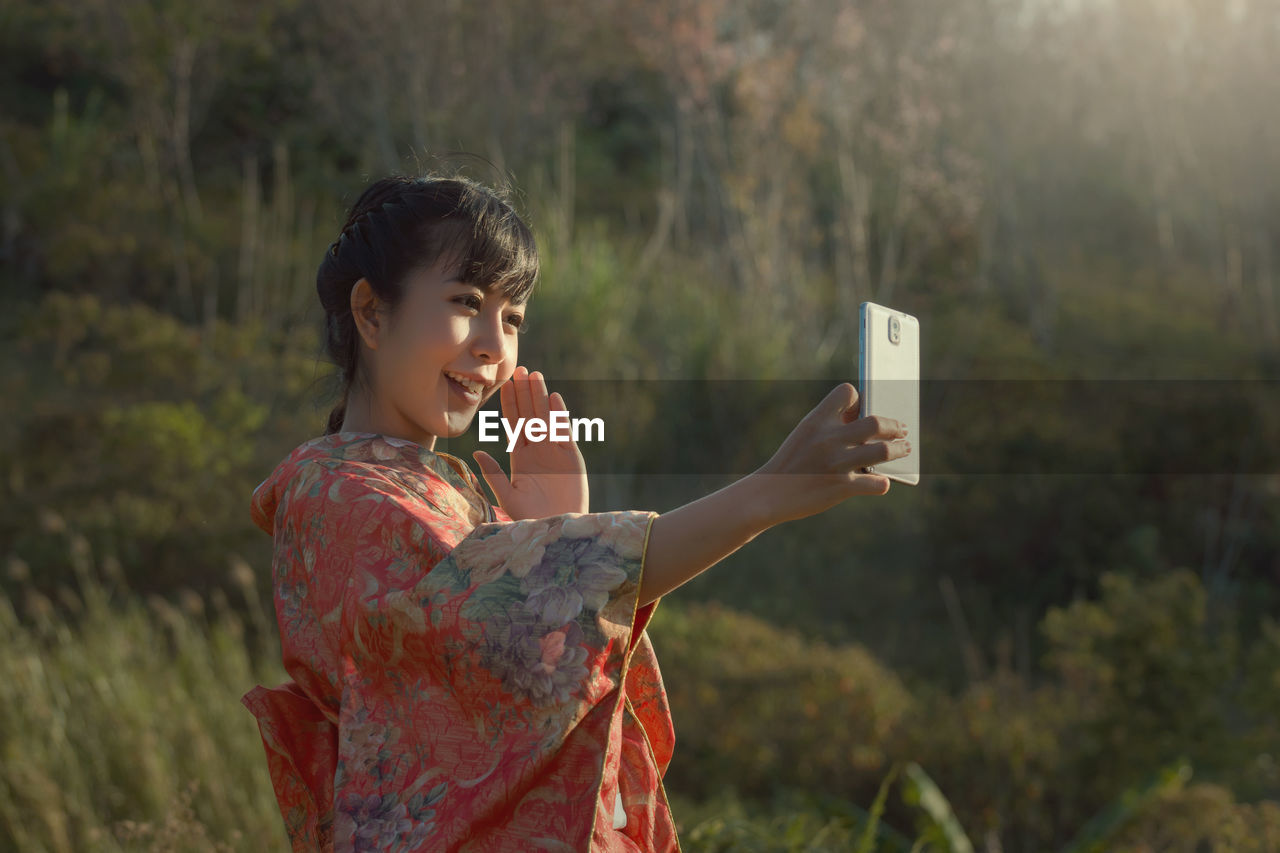 Close-up of smiling woman taking selfie through smart phone against trees