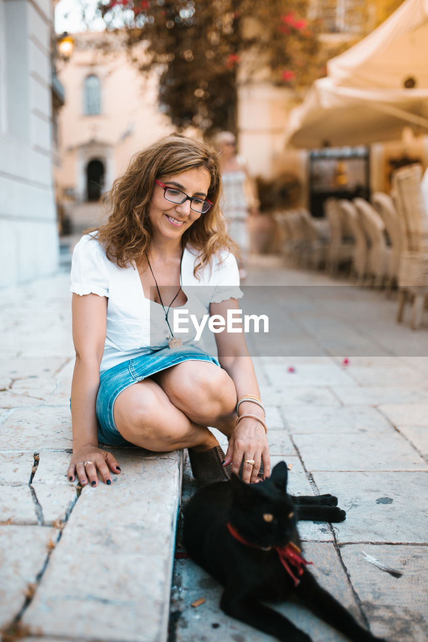 FULL LENGTH OF WOMAN SITTING ON SIDEWALK IN CITY