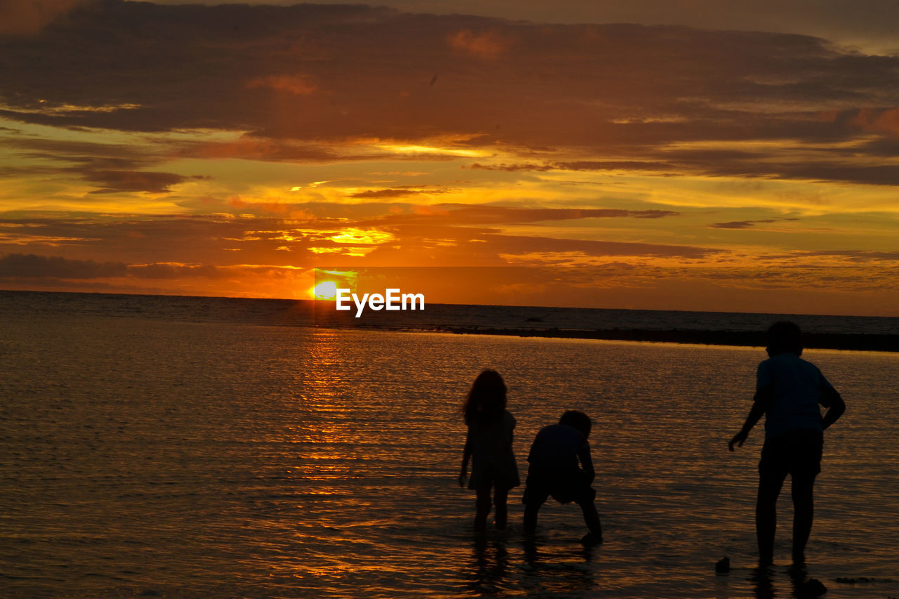 Silhouette children playing in sea against sky during sunset