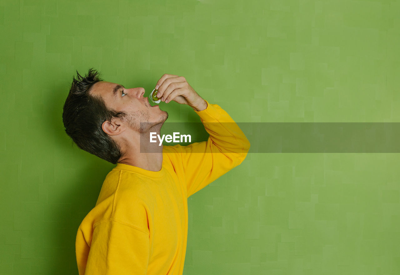 Young man drinking olive oil in a small plastic ampoule on a green background, healthy lifestyle