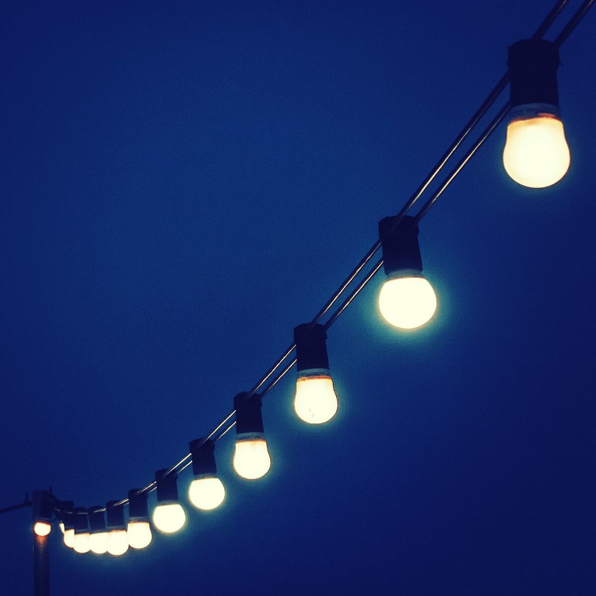 Low angle view of illuminated light bulbs on cable against clear blue sky at dusk