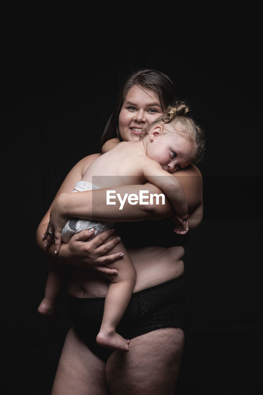 Young plus size woman in underwear holding cute little child in arms and looking at camera with happy smile while standing against black background