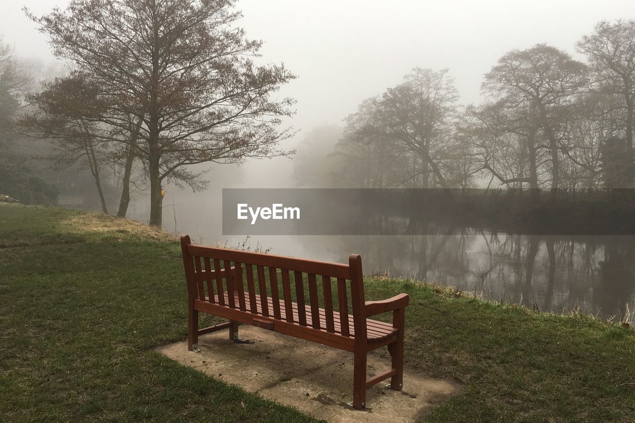 Empty bench by calm lake in foggy weather