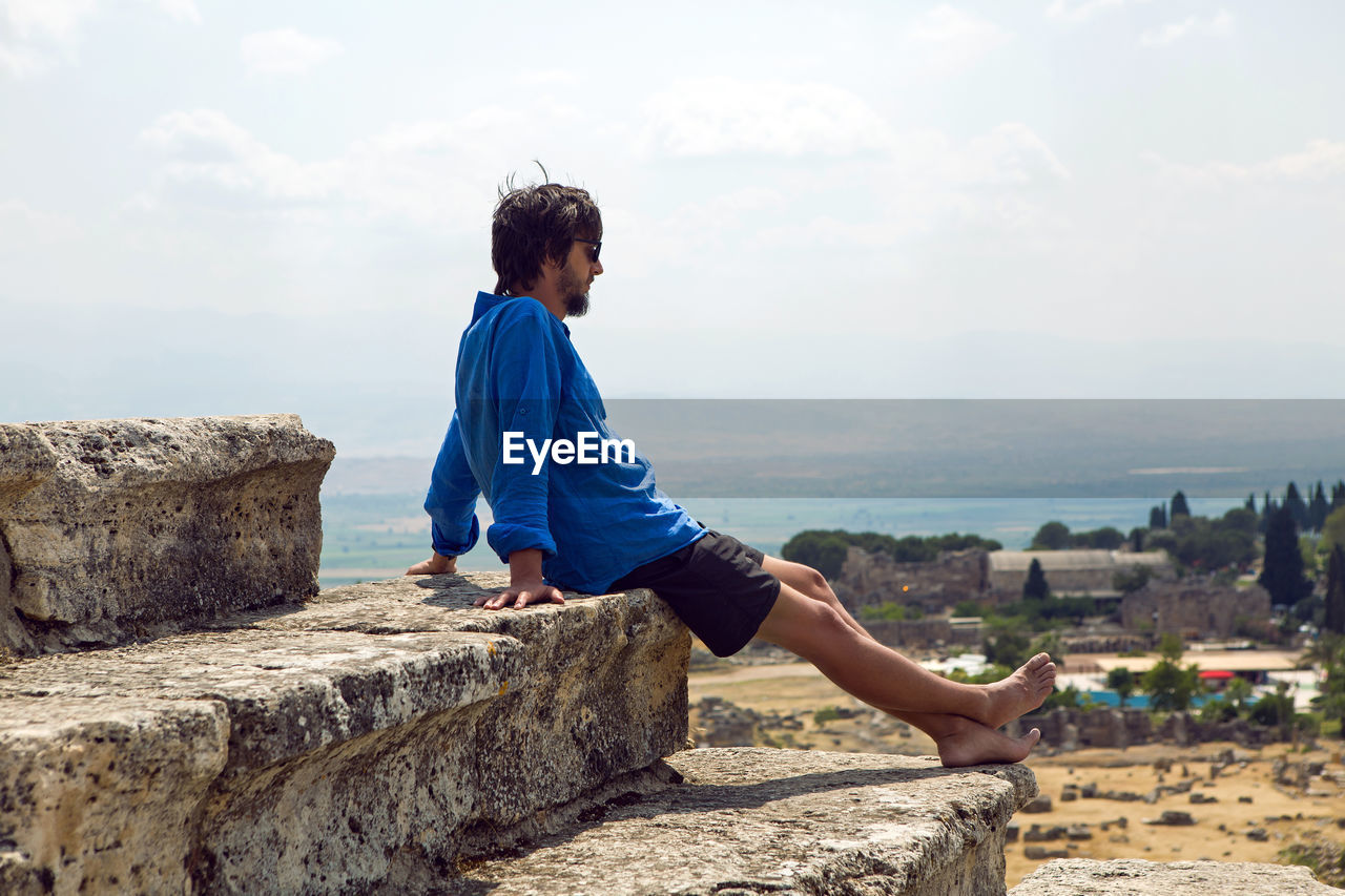 Man in a blue linen shirt and shorts sits on an old stone staircase colosseum in pamukkale turkey
