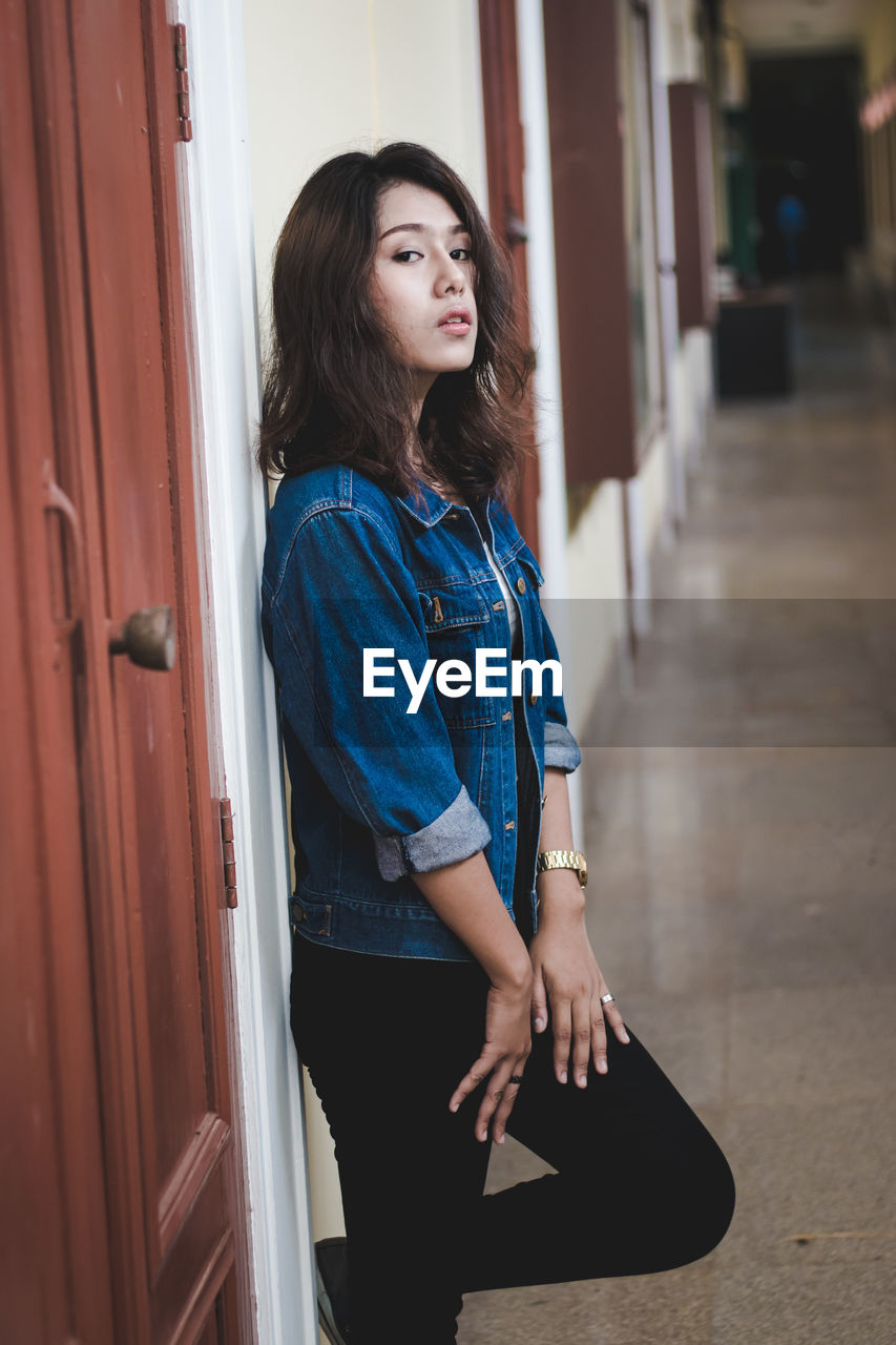 Portrait of confident young woman leaning on wall while standing in corridor