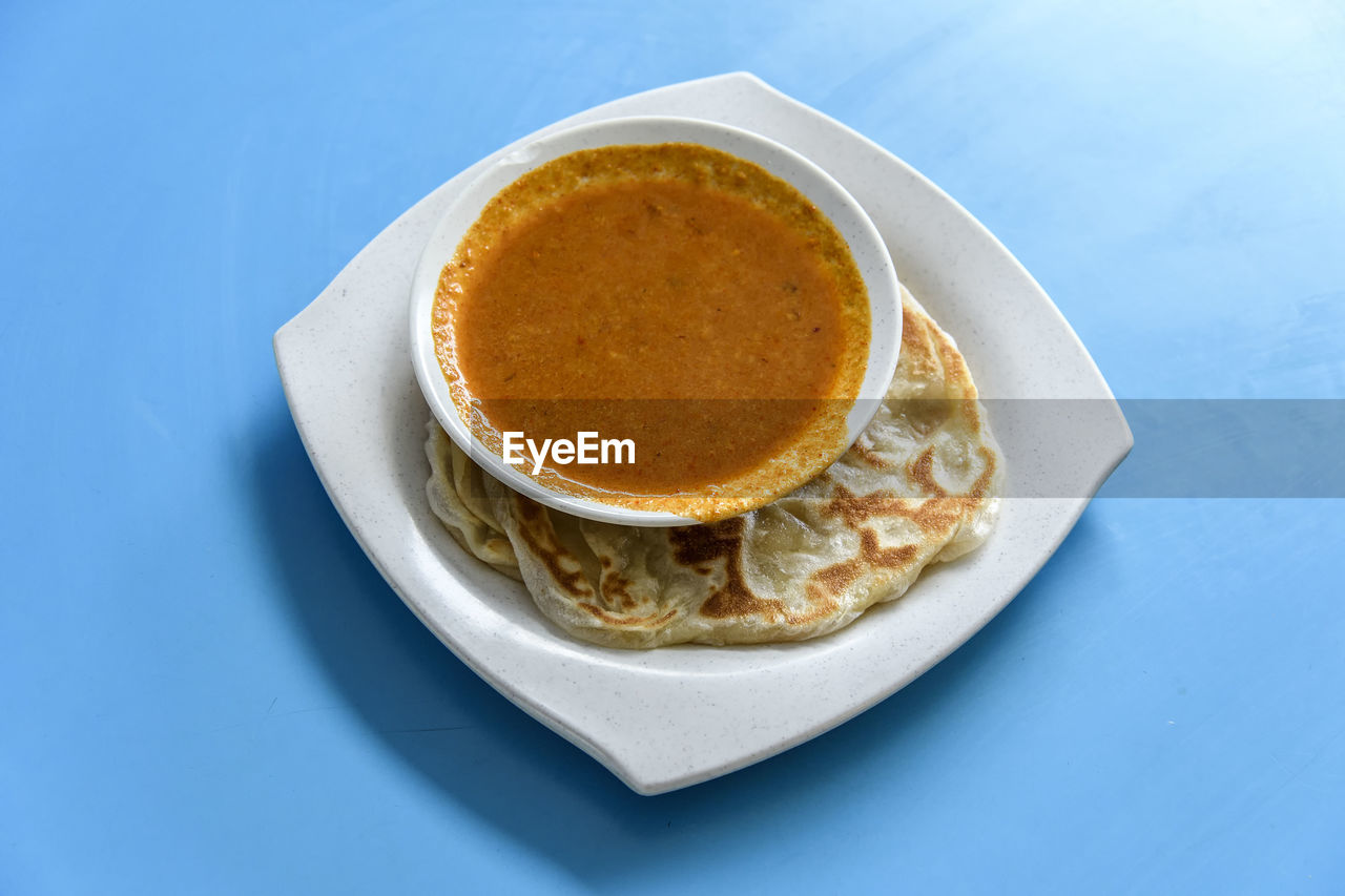 High angle view of fresh roti prata served with curry in plate on blue table