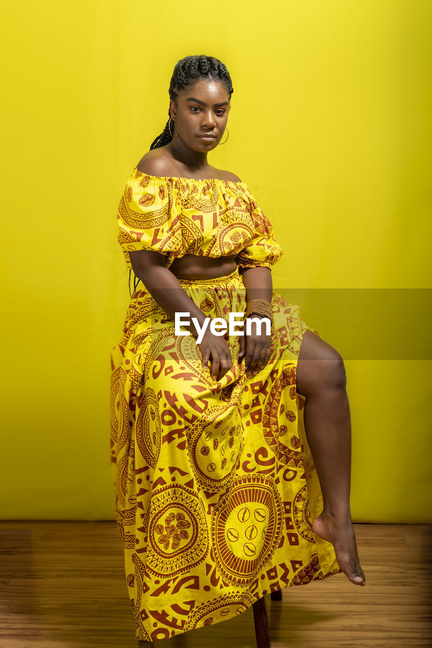 Portrait of young, beautiful woman sitting on a wooden stool. against yellow background.