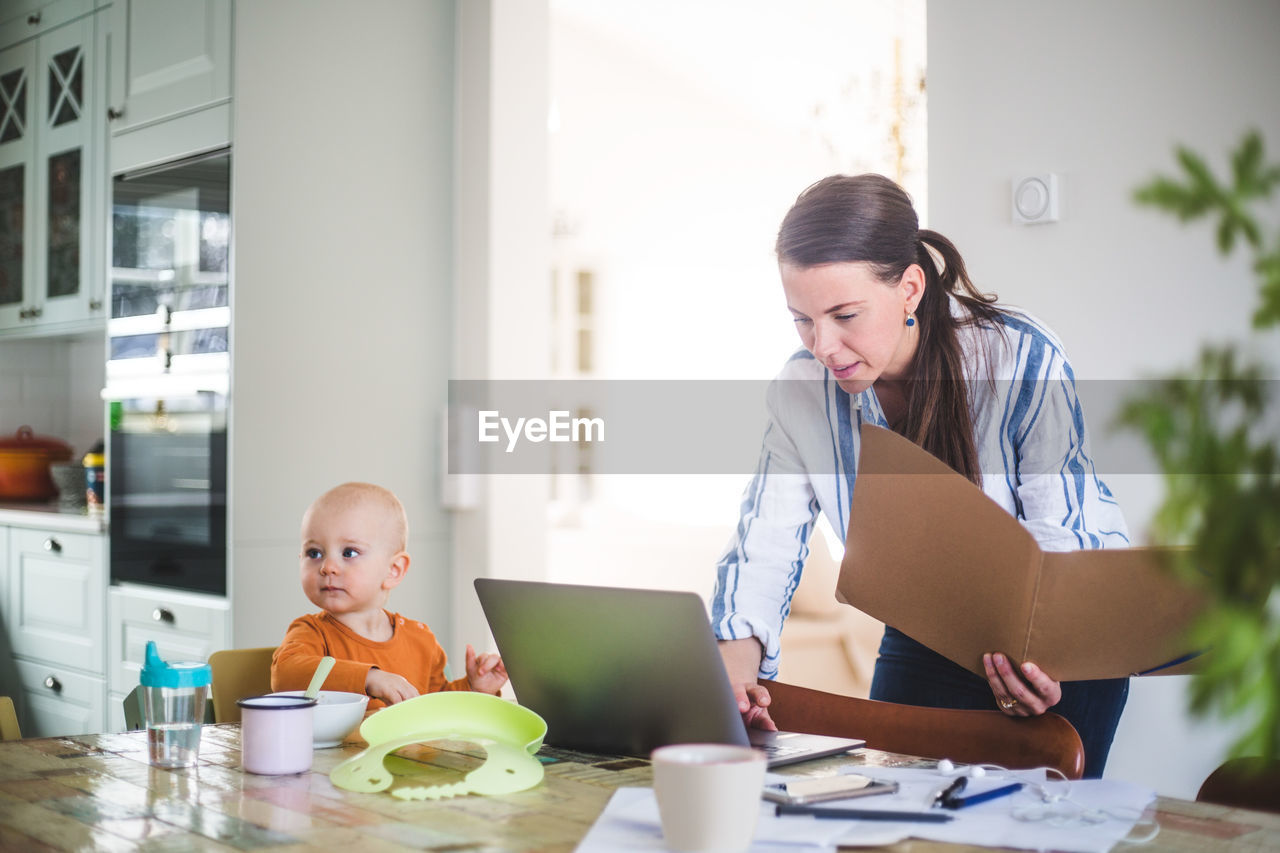 Female professional holding file while using laptop by daughter on dining table at home office