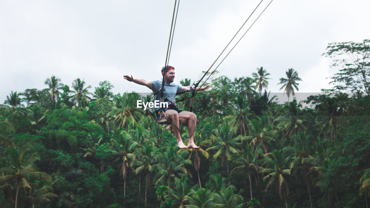 Full length of young man sitting on rope swing against trees and sky