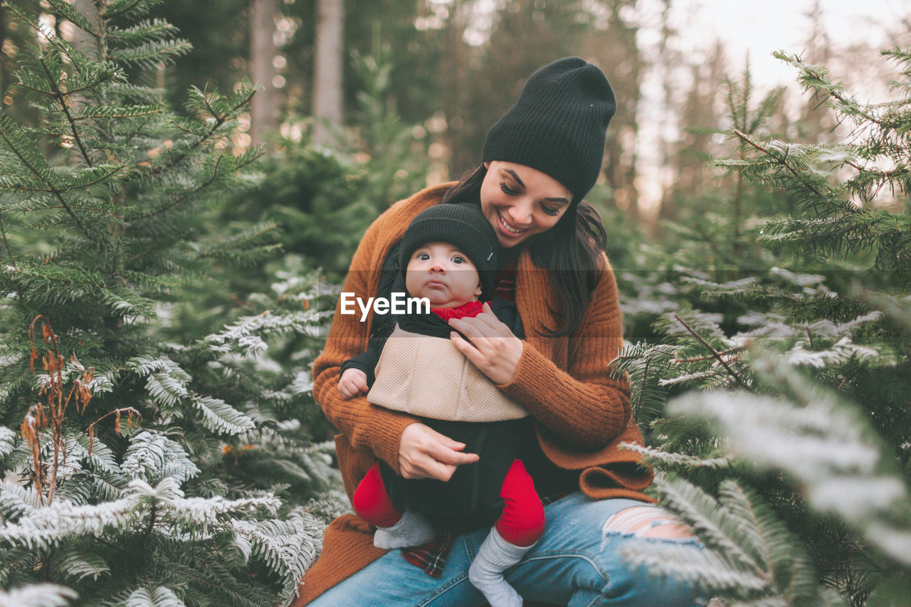 Mother with cute son crouching amidst pine trees in forest during winter