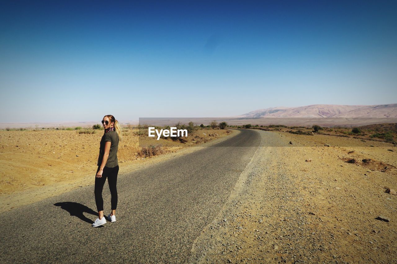 Portrait of mid adult woman standing on road against clear blue sky