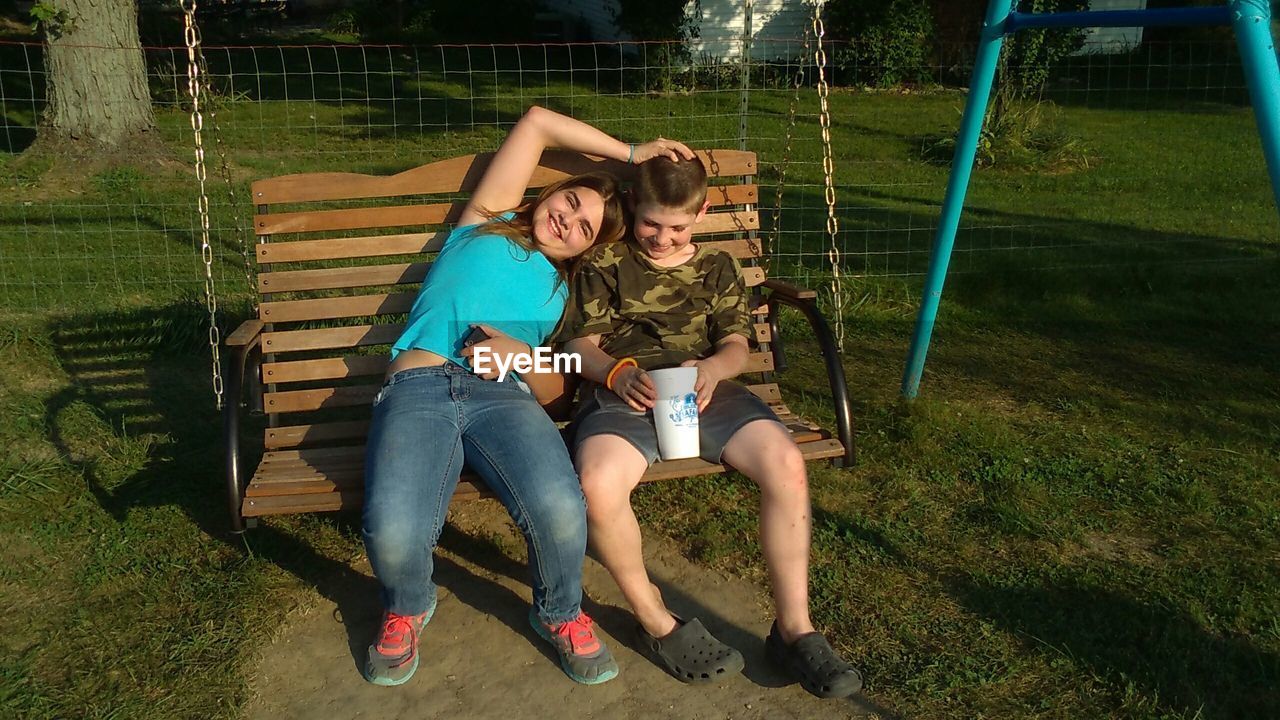 Brother and sister sitting on swing at grassy field