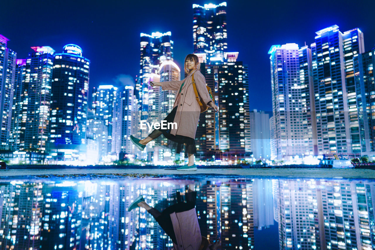 Portrait of woman standing by lake against illuminated buildings in city at night