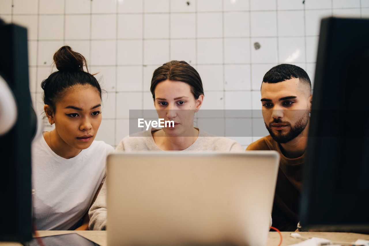 Young multi-ethnic computer hackers looking at laptop against wall in office