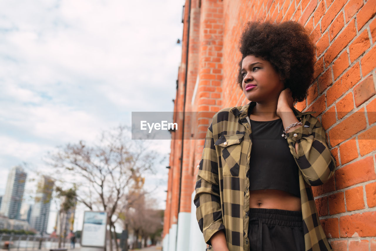 YOUNG WOMAN LOOKING AWAY WHILE STANDING IN FRONT OF TRADITIONAL BUILDING